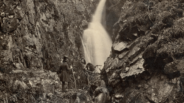 A black and white vintage photograph of a waterfall cascading through a gully with a man in a tall black hat standing looking at it